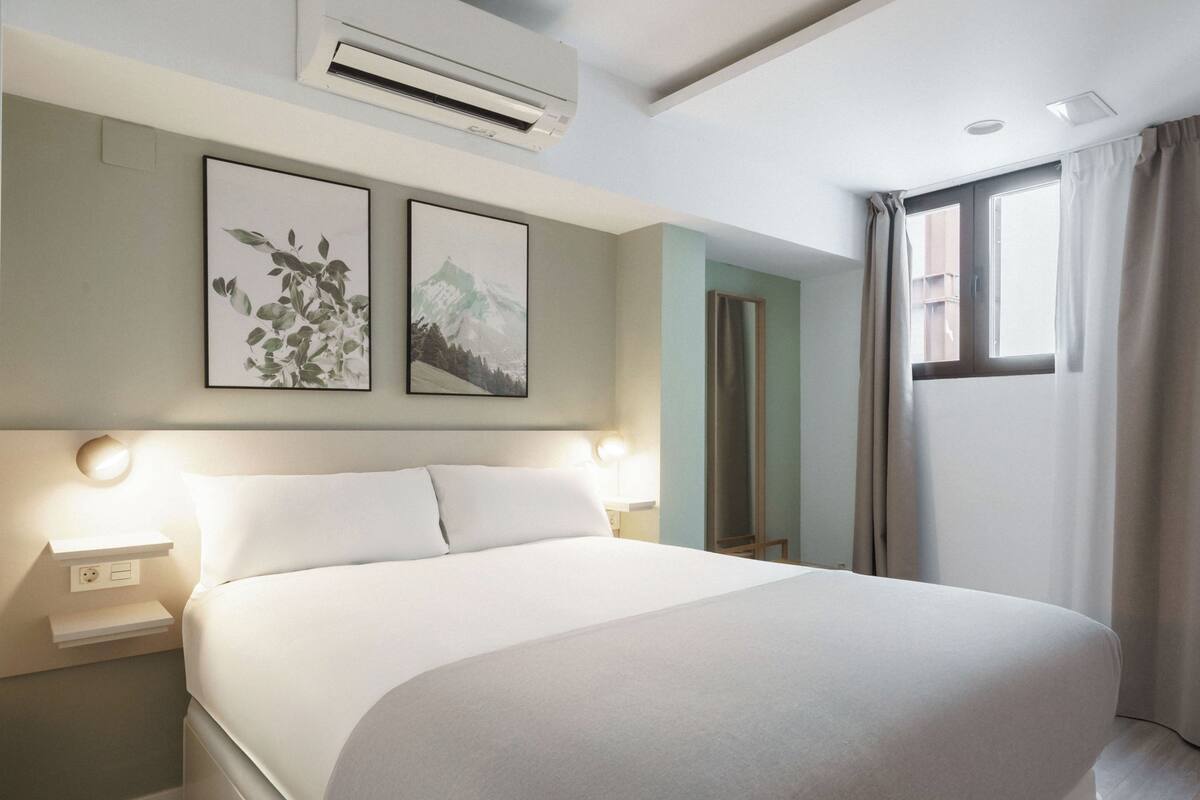 Habitación de hotel con una cama grande, una mesita de noche con un teléfono, un aire acondicionado y dos cuadros en la pared.
