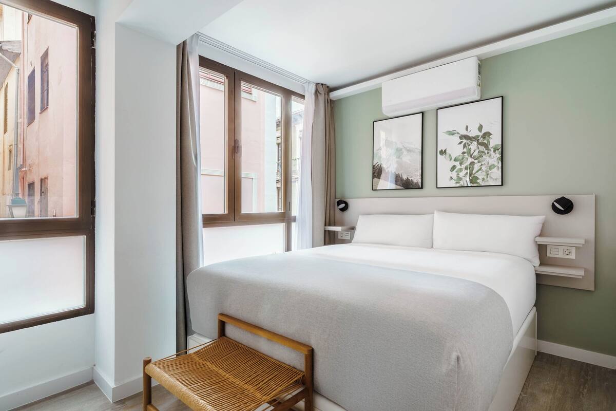 Una habitación de hotel moderna con una cama grande, una pequeña silla de madera, una mesita de noche y un ventanal con cortinas.