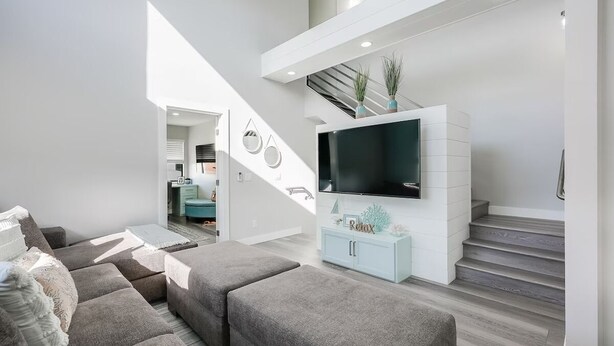 A modern living room with a grey sectional sofa, a flat-screen TV on a white console, and a staircase leading to an upper level. - Luxury Meets Coastal Charm in North Mission Beach (San Diego)