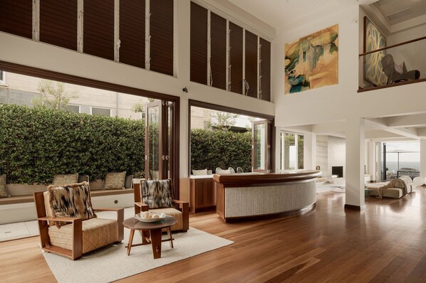 A spacious living area with a high ceiling, a large window, and a view of a green hedge outside.