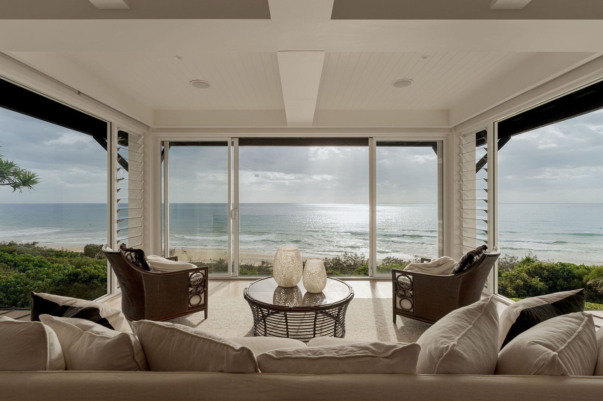 A room with a large window overlooking the ocean, furnished with a sofa and two wicker chairs, and a small round table with decorative items.