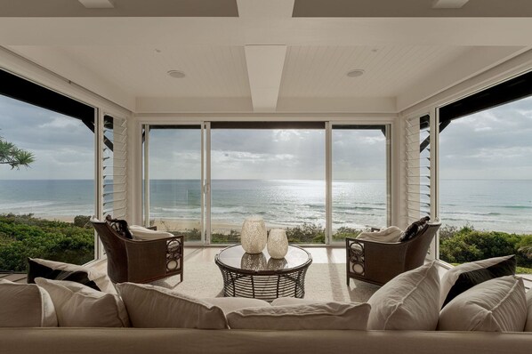 A room with a large window overlooking the ocean, furnished with a sofa and two wicker chairs, and a small round table with decorative items.