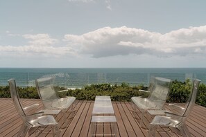 A deck with clear plastic chairs and a table overlooking the ocean.