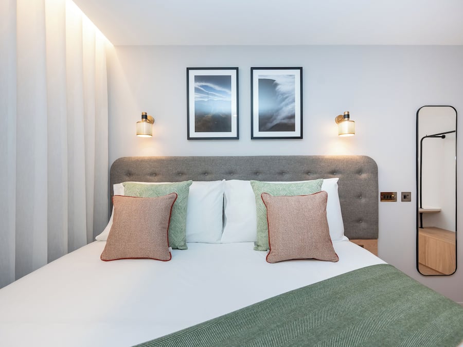 Un lit bien fait, avec une tête de lit grise, deux tableaux encadrés au mur et un miroir sur le côté droit.