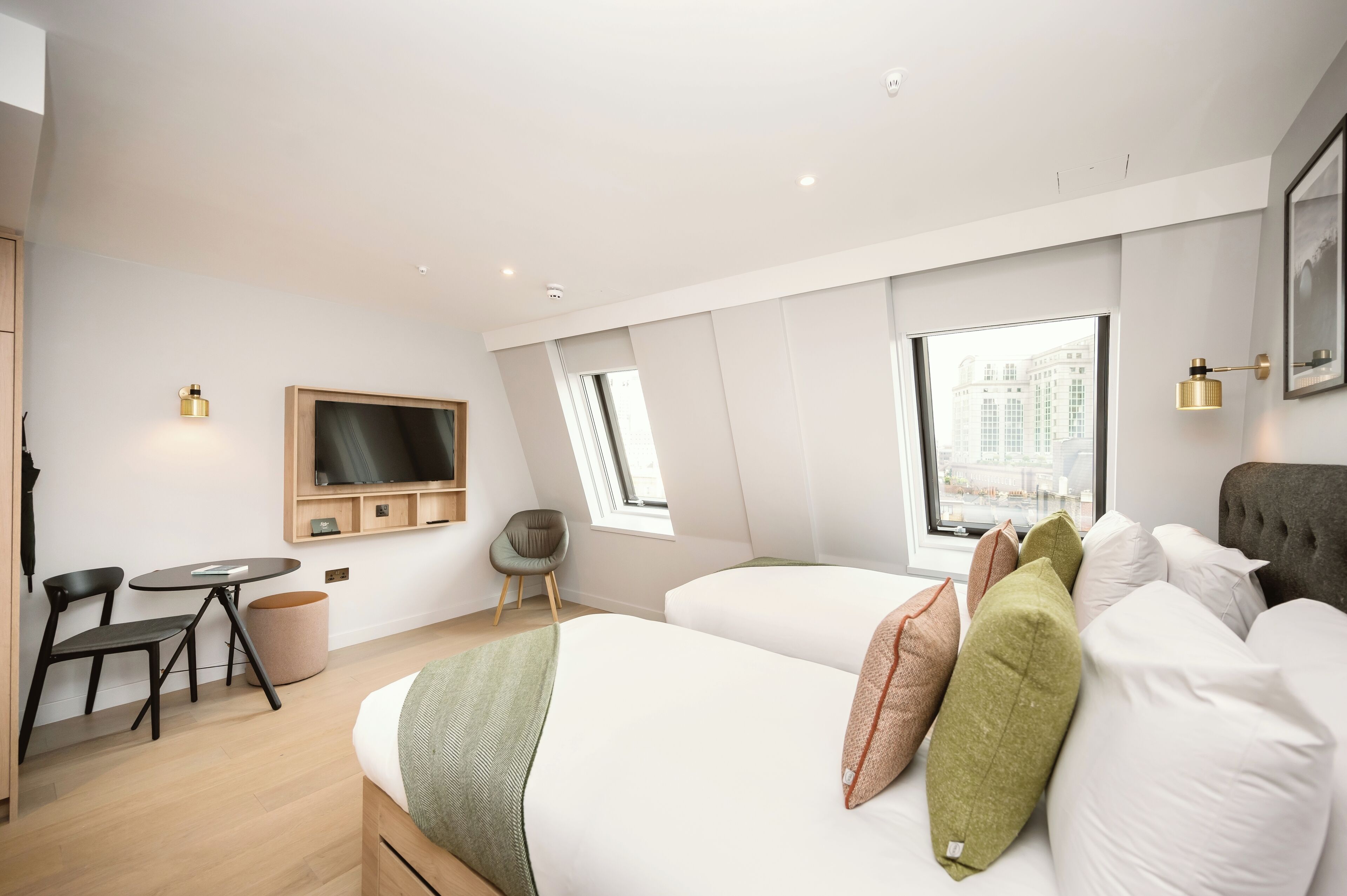 A modern hotel room with a bed, a desk, a chair, a TV mounted on the wall, and large windows offering a city view.