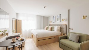 Een moderne slaapkamer met een bed, een groene fauteuil, een eettafel met een schaal fruit en een raam met dunne gordijnen.