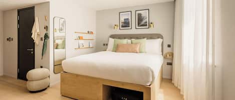 A modern bedroom with a large bed, a wooden nightstand, a mirror, and a shelf with toiletries.