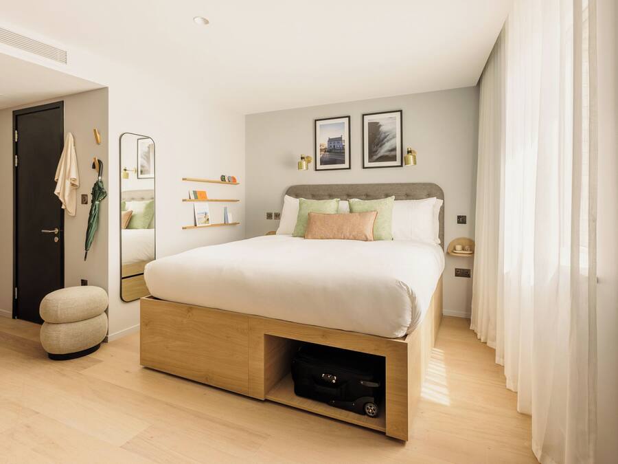 A modern bedroom with a large bed, a wooden nightstand, a mirror, and a shelf with toiletries.