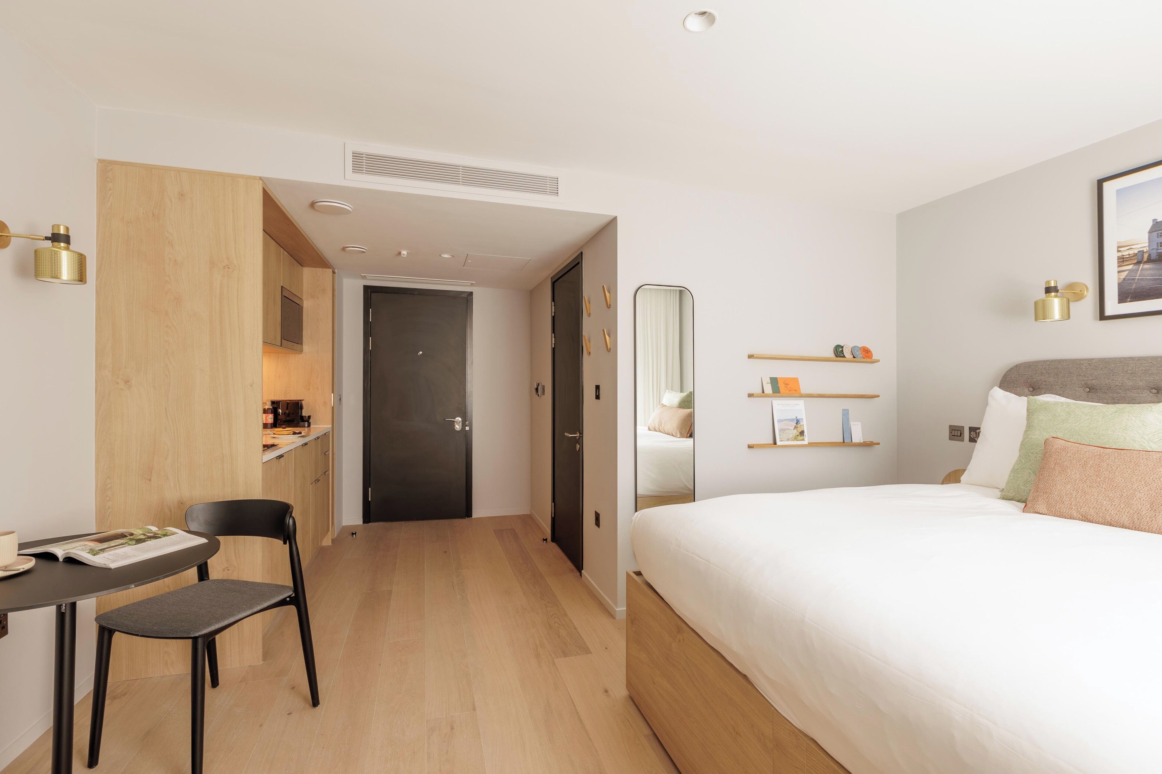A modern hotel room with a wooden floor, a bed with white bedding, a desk with a chair, a small kitchen area, and a large mirror.