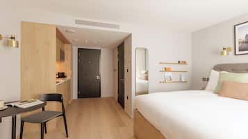 Une chambre d’hôtel moderne avec un plancher en bois, un lit avec une literie blanche, un bureau avec une chaise, un petit coin cuisine et un grand miroir.