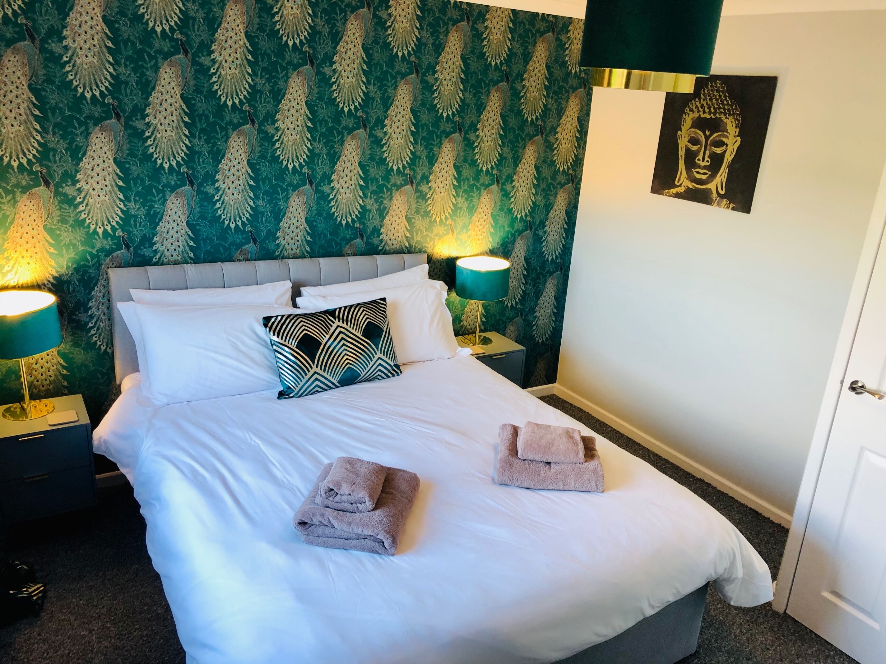 Un lit bien fait, avec du linge de lit blanc, deux oreillers et deux serviettes pliées. On y trouve deux lampes de chevet avec des abat-jour turquoise, et un mur recouvert d’un papier peint à motifs de paon. Une statue de Bouddha est visible sur le mur.