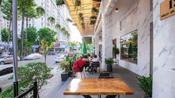 Un café al aire libre con una mesa y sillas de madera, rodeado de plantas en macetas y vistas a una calle de la ciudad.