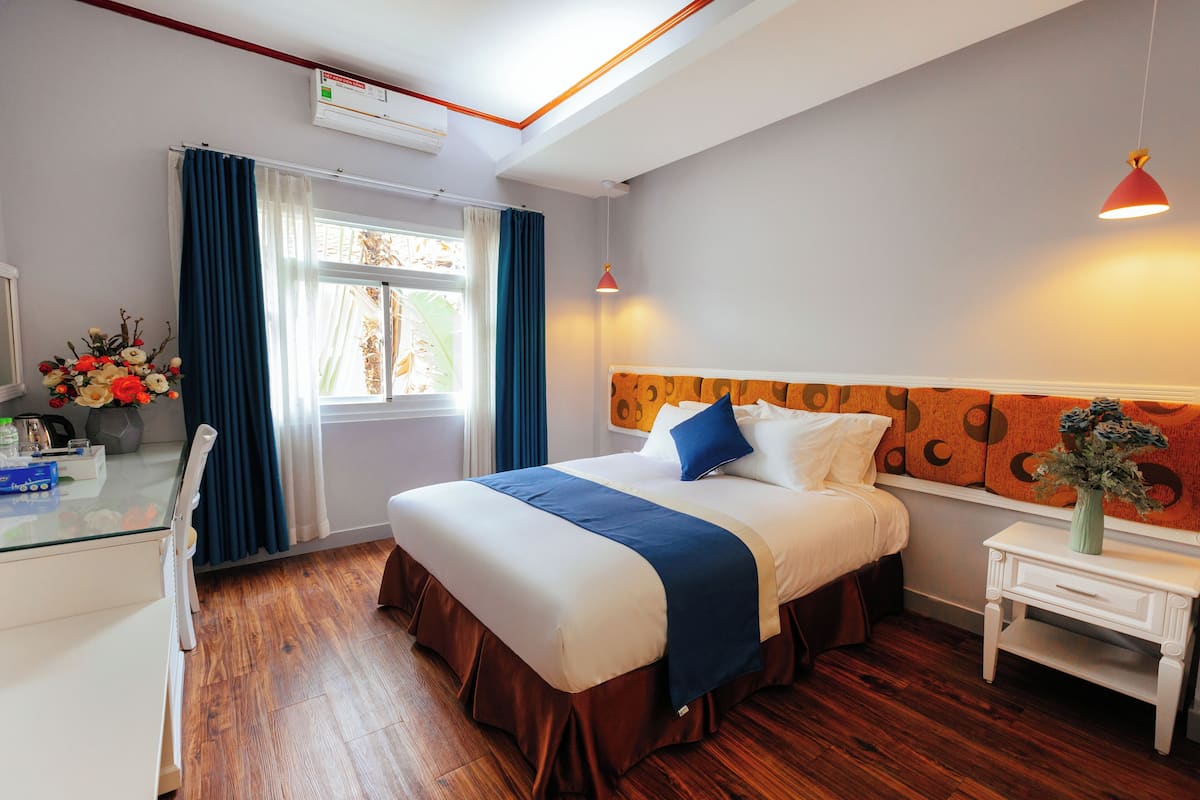 Quarto de hotel com cama, escrivaninha, cadeira, janela com cortinas, mesa de cabeceira e ar-condicionado fixado na parede.