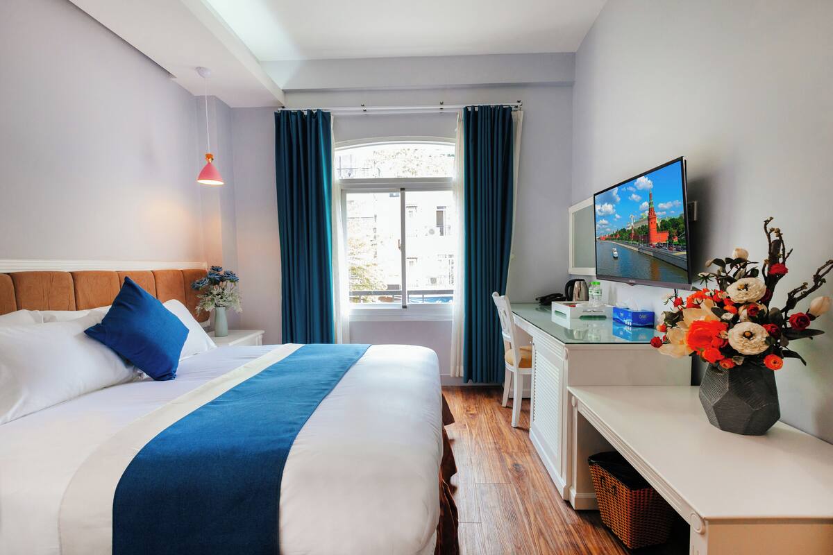 Quarto de hotel com cama, escrivaninha, televisão e janela com cortinas.