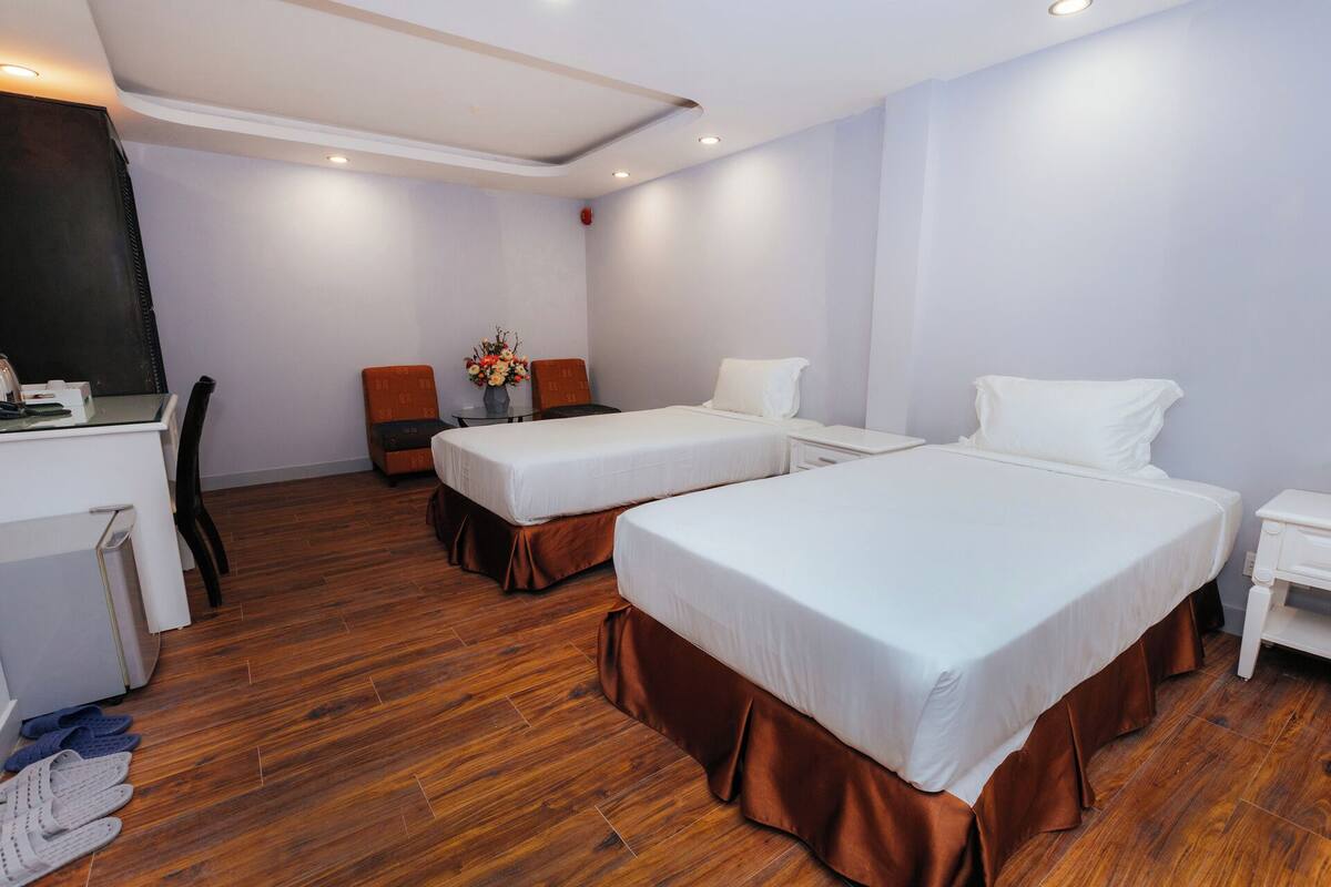 Quarto de hotel com duas camas, uma escrivaninha, uma cadeira e uma mesinha com um vaso de flores.