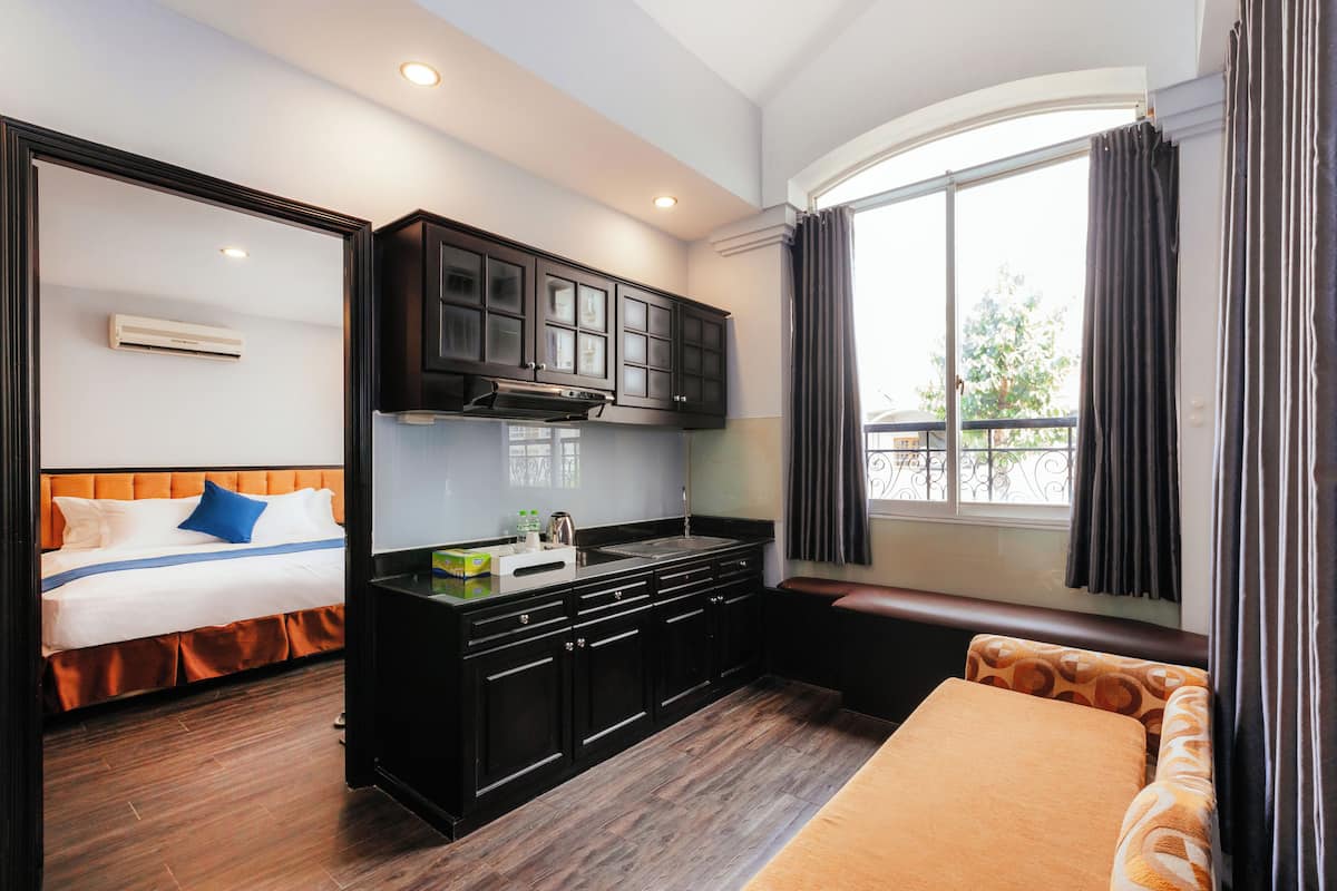 Quarto de hotel compacto com cozinha compacta, cama com roupa de cama laranja e uma janela grande com cortinas.