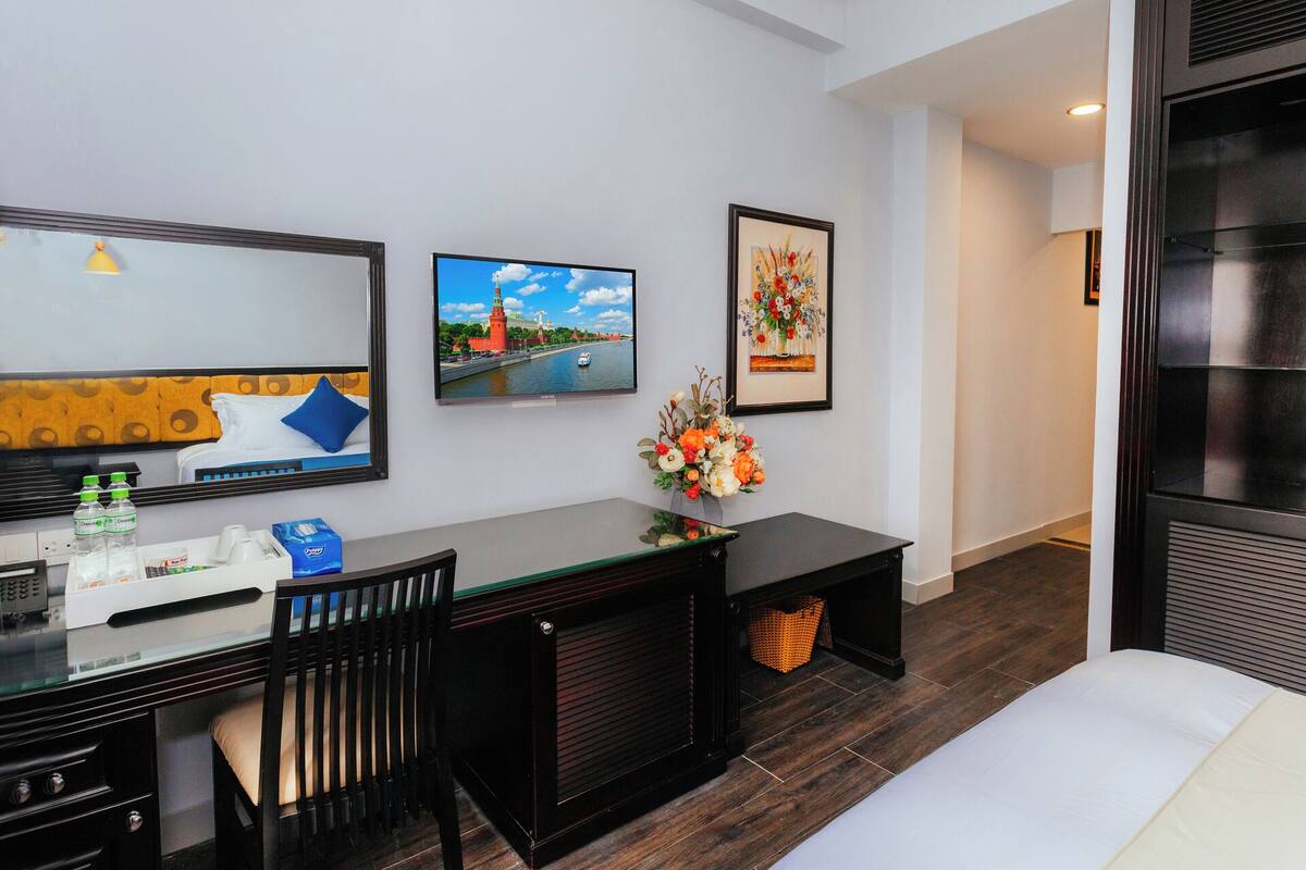 Quarto de hotel com uma cama, uma escrivaninha com um computador, uma cadeira, uma televisão e quadros nas paredes.