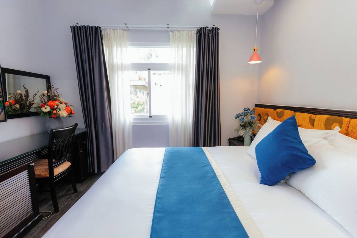 Quarto de hotel com cama, escrivaninha, cadeira, janela com cortinas e luminária pendente.