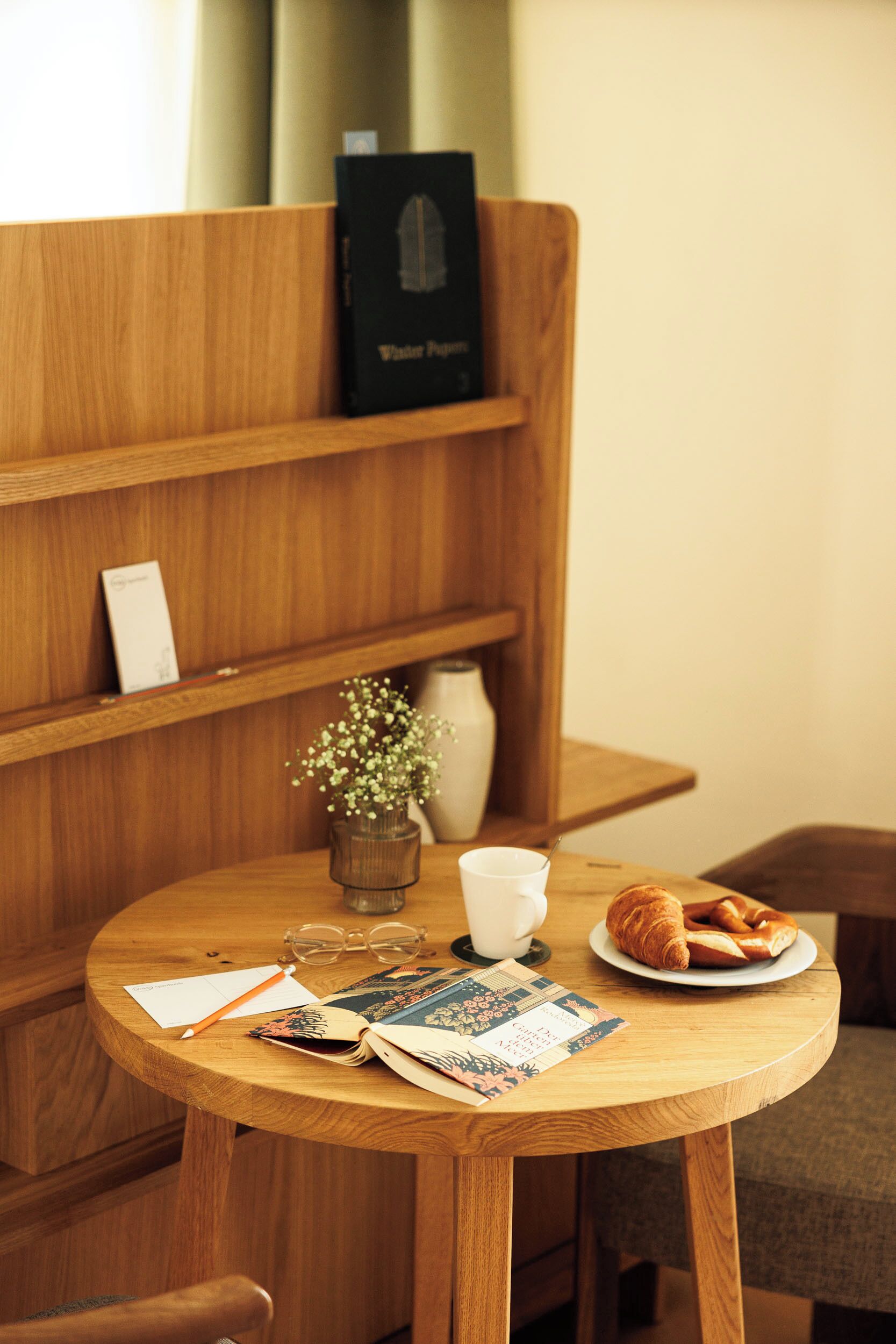 Ein Holztisch mit einem Buch, einer Tasse, einem Brillenpaar, einer kleinen Vase mit Blumen und einem Croissant.
