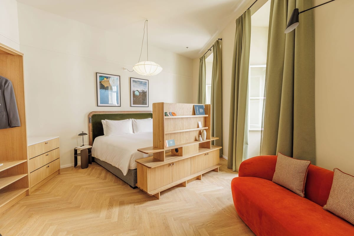 A bedroom with a bed, a wooden nightstand, a bedside table, a bookshelf, a red sofa, and a window with curtains.