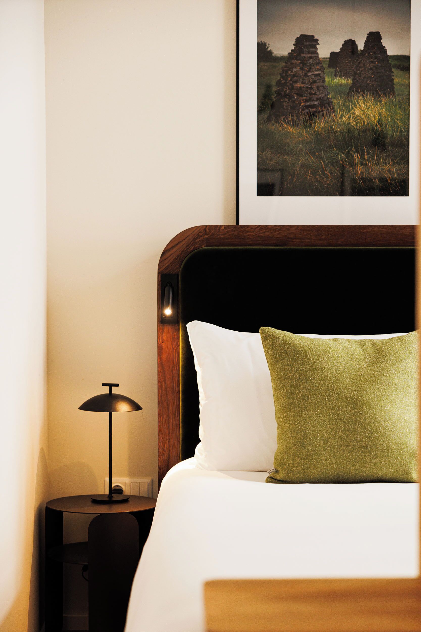 A neatly made bed with white and green pillows, a wooden headboard, and a framed picture of ruins on the wall.