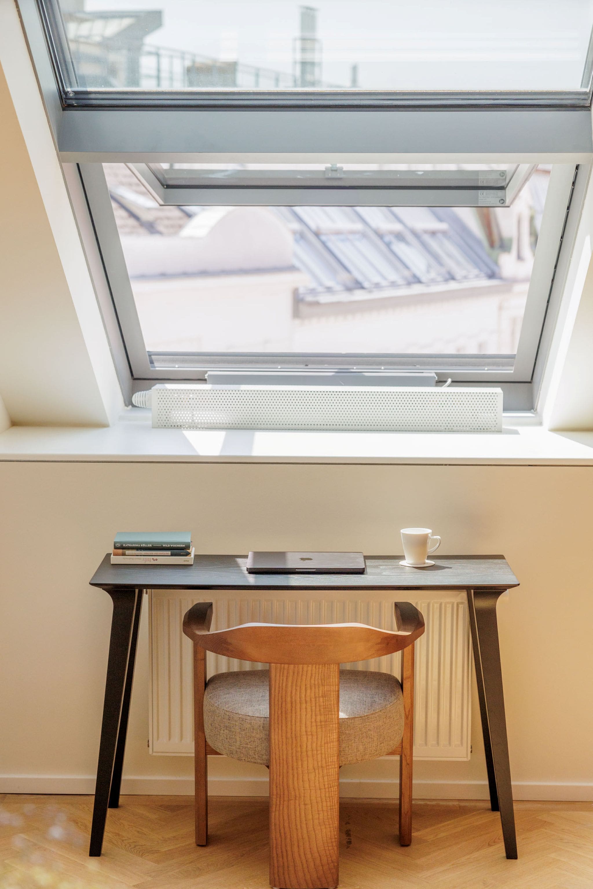 Une fenêtre de toit, un radiateur, une chaise en bois, une petite table avec un livre et une tasse, et un ordinateur portable.