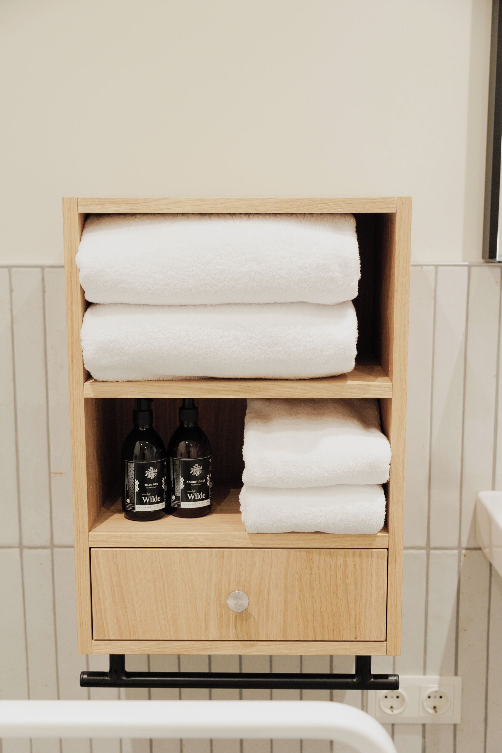 Une étagère en bois avec des serviettes blanches pliées et deux bouteilles de gel douche de la marque Wilde.