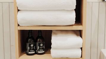 Une étagère en bois avec des serviettes blanches pliées et deux bouteilles de gel douche de la marque Wilde.