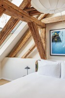Quarto com teto inclinado, vigas de madeira, uma cama, um quadro emoldurado e um abajur.