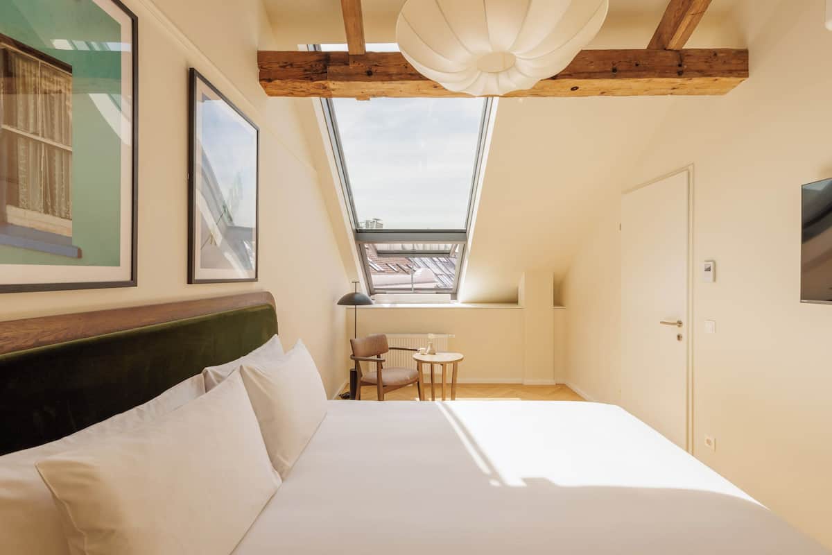 A bedroom with a skylight, a bed, a chair, a small table, and a TV on the wall.