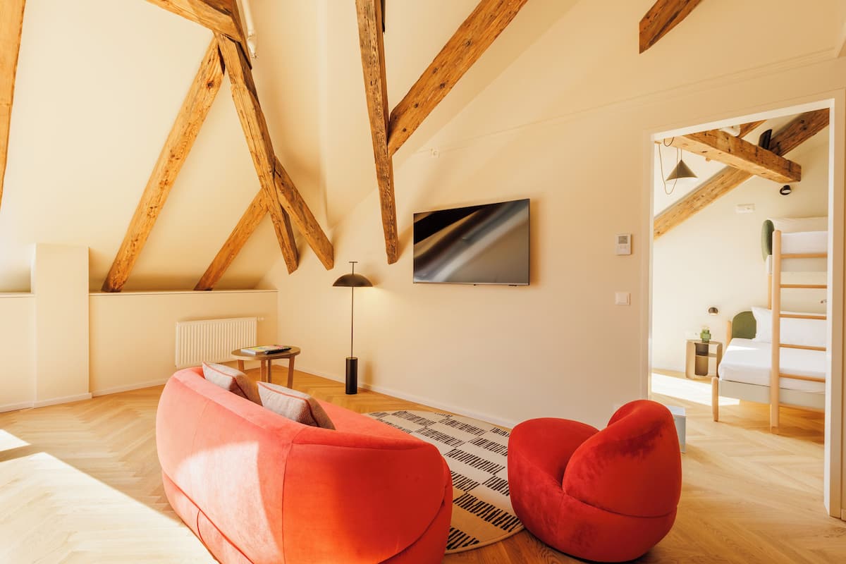 A modern living room with exposed wooden beams, a red sofa, a floor lamp, and a view into a bedroom with a bunk bed.