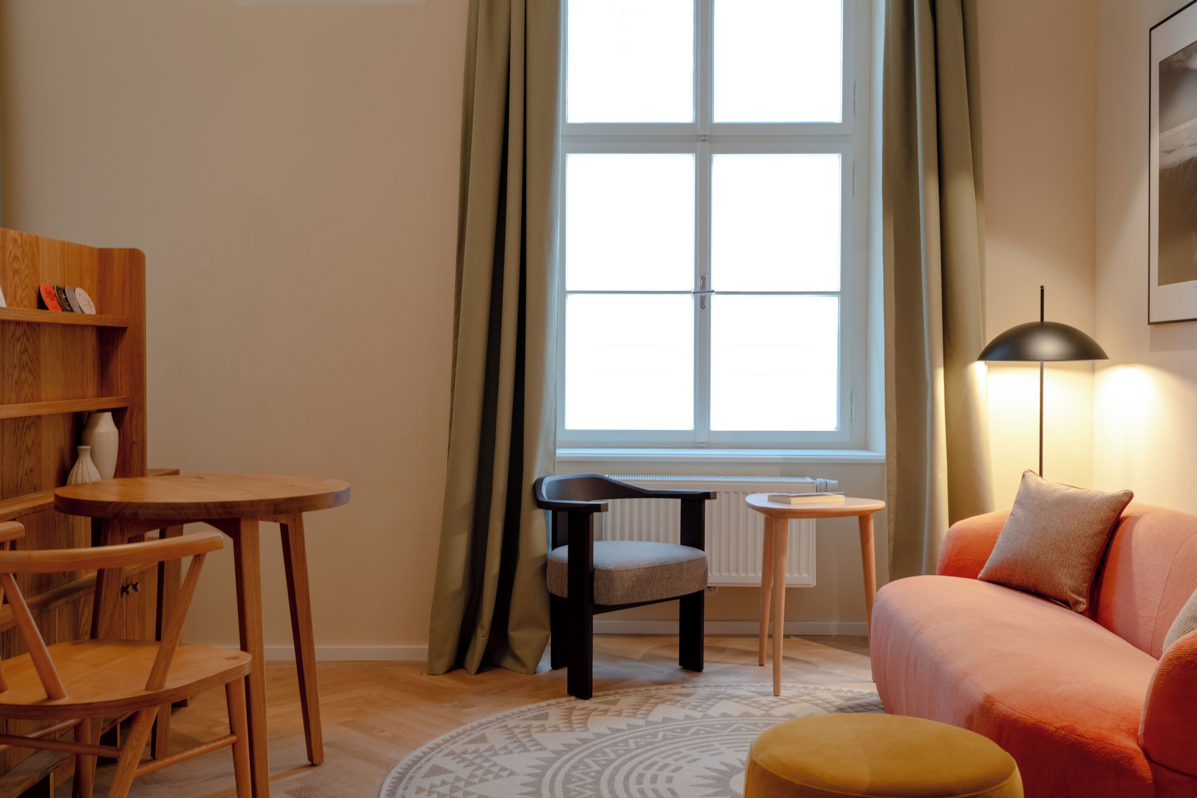 A cozy living room with a sofa, a small table, and a chair. There is a window with curtains, a radiator, and a lamp.