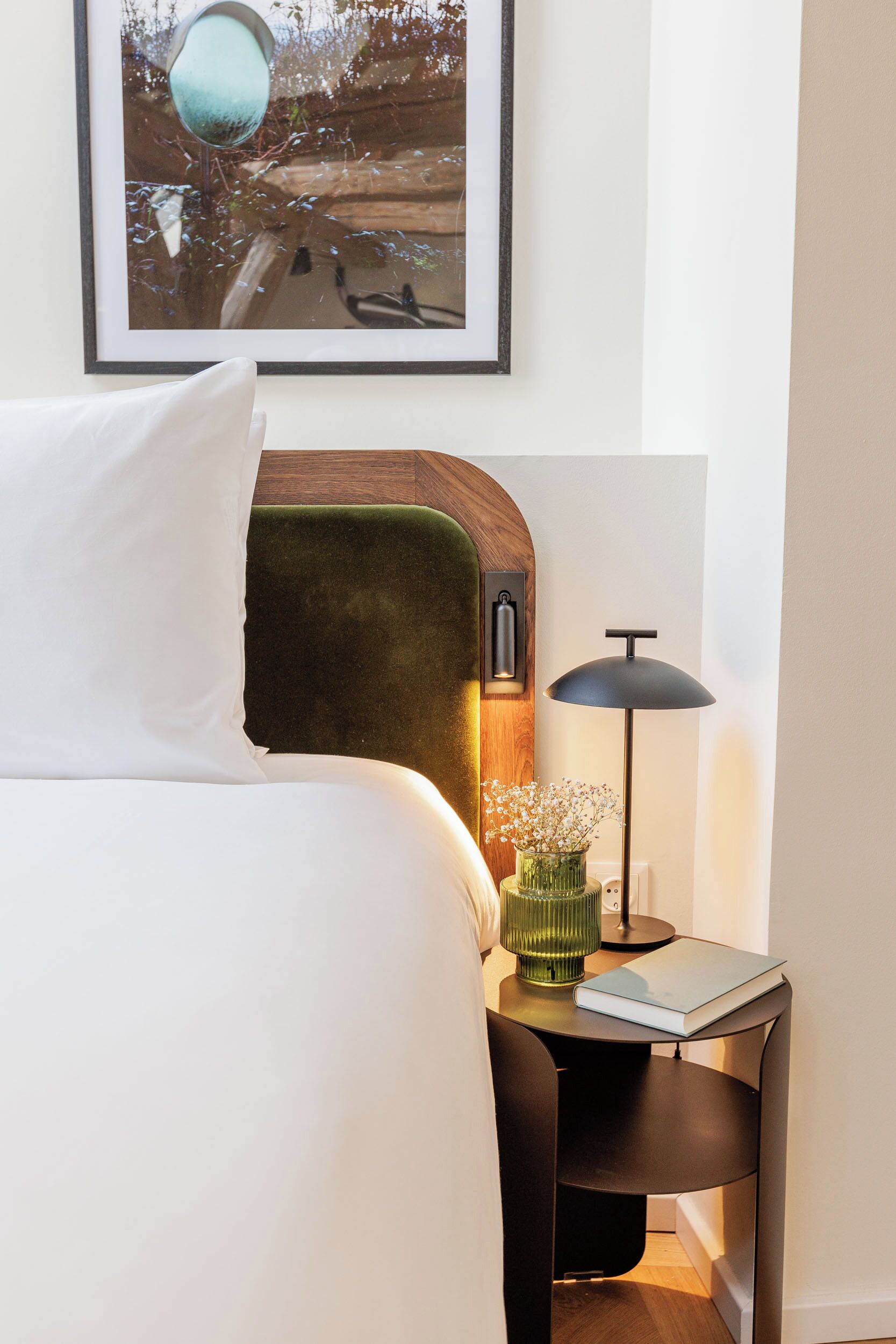 A neatly made bed with a pillow, a bedside table with a lamp, a framed picture on the wall, and a small vase with flowers.