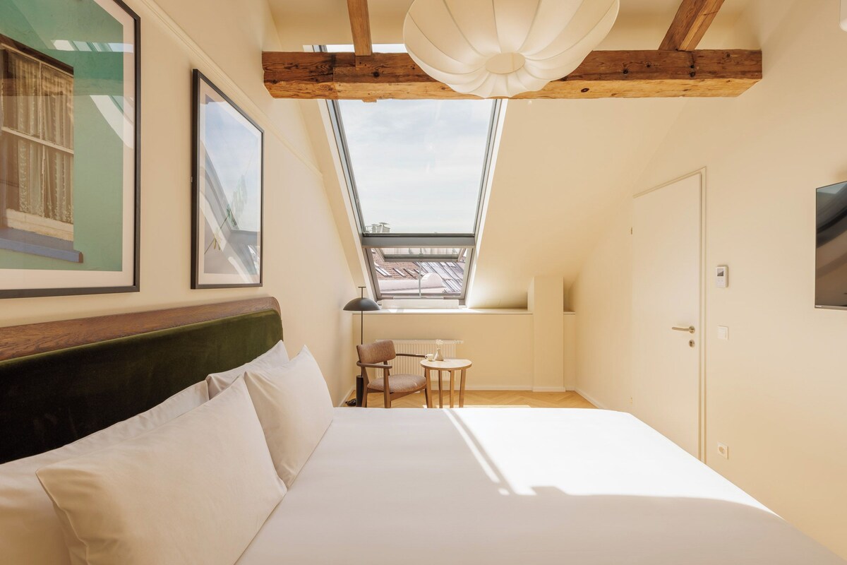 Un dormitorio con tragaluz, una cama, una silla, una mesita pequeña y un televisor en la pared.