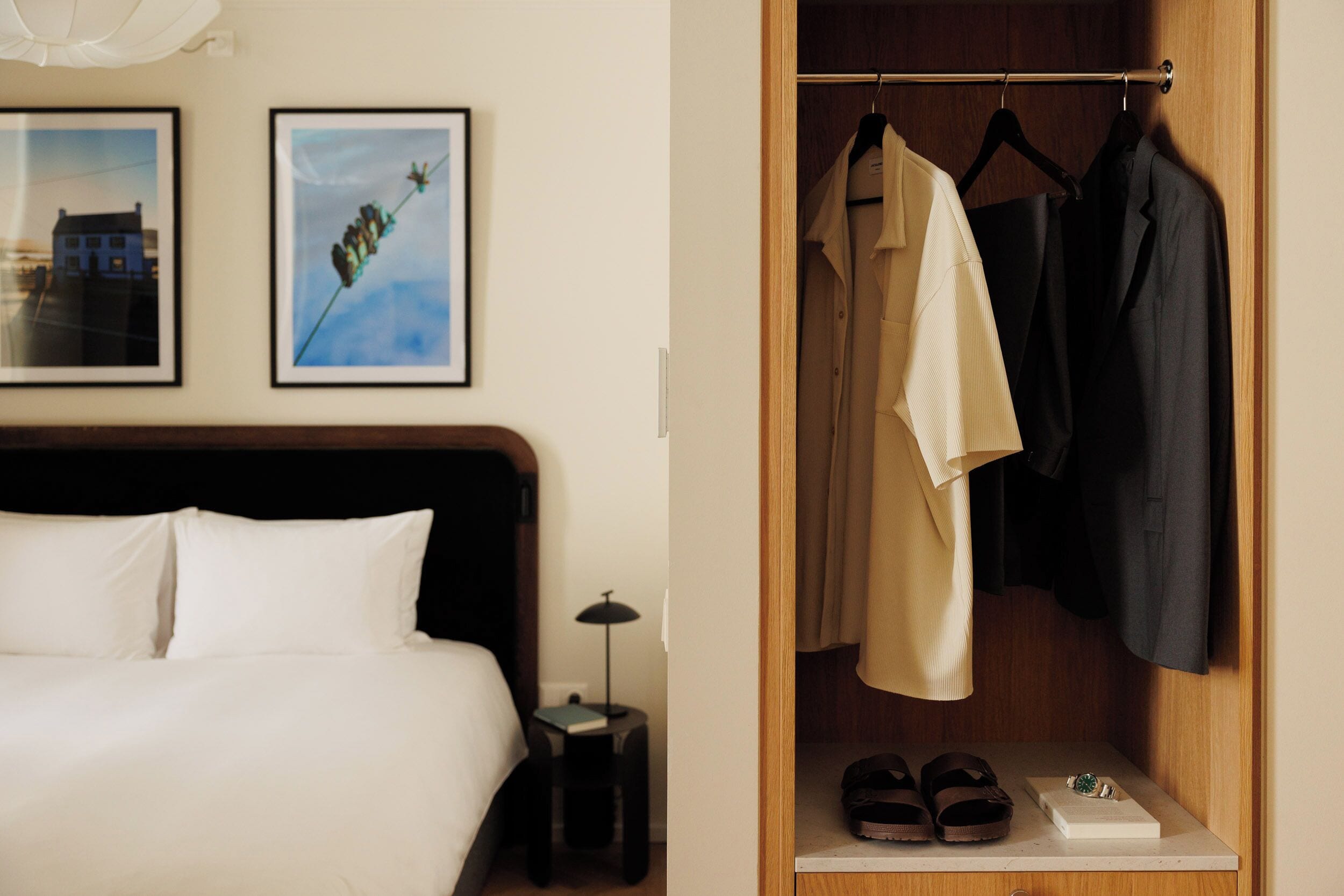 A hotel room with a bed, a nightstand with a lamp, a closet with hanging clothes, and a shelf with shoes and a box.