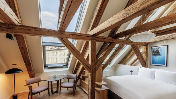 Ein Schlafzimmer mit einem großen Bett, Holzbalken, einem Oberlicht, einem Schreibtisch mit Stühlen und Blick auf ein Gebäude.