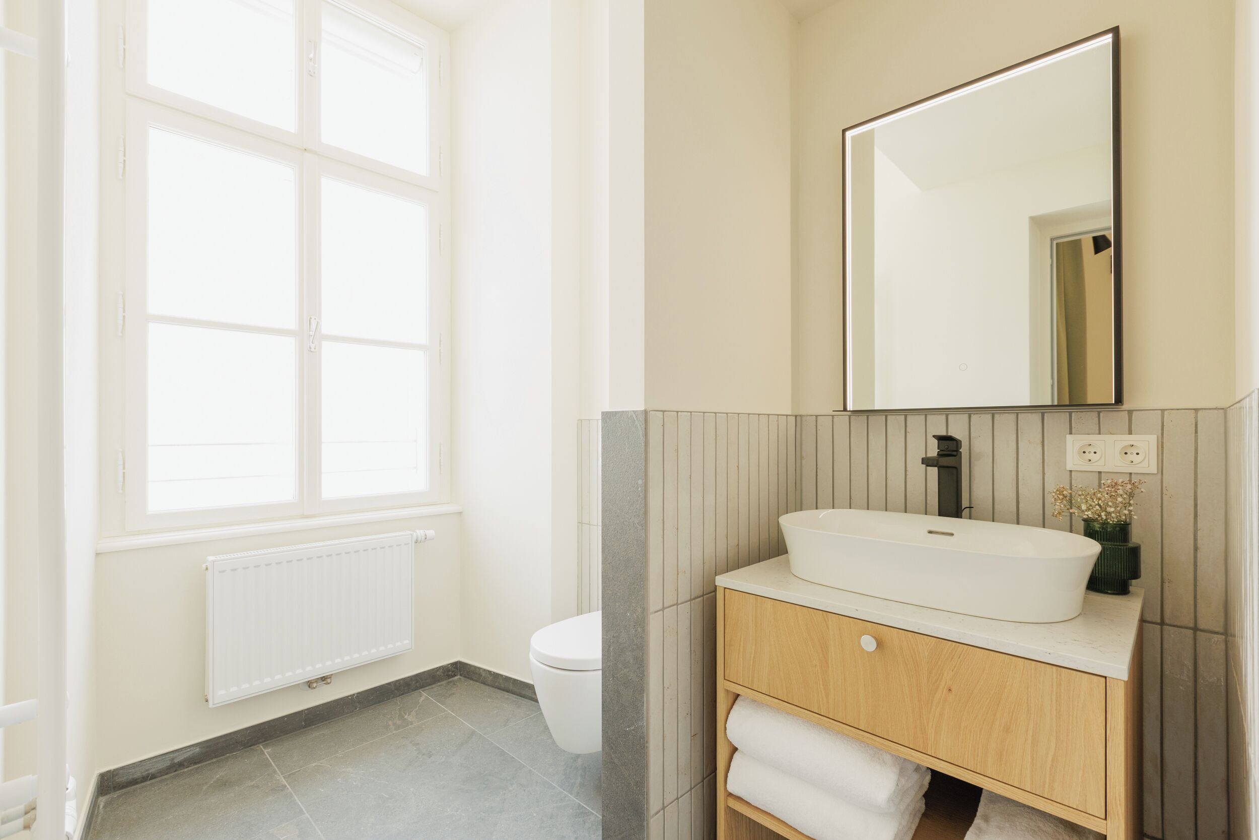 A bathroom with a large window, a white sink, a mirror, and a toilet.