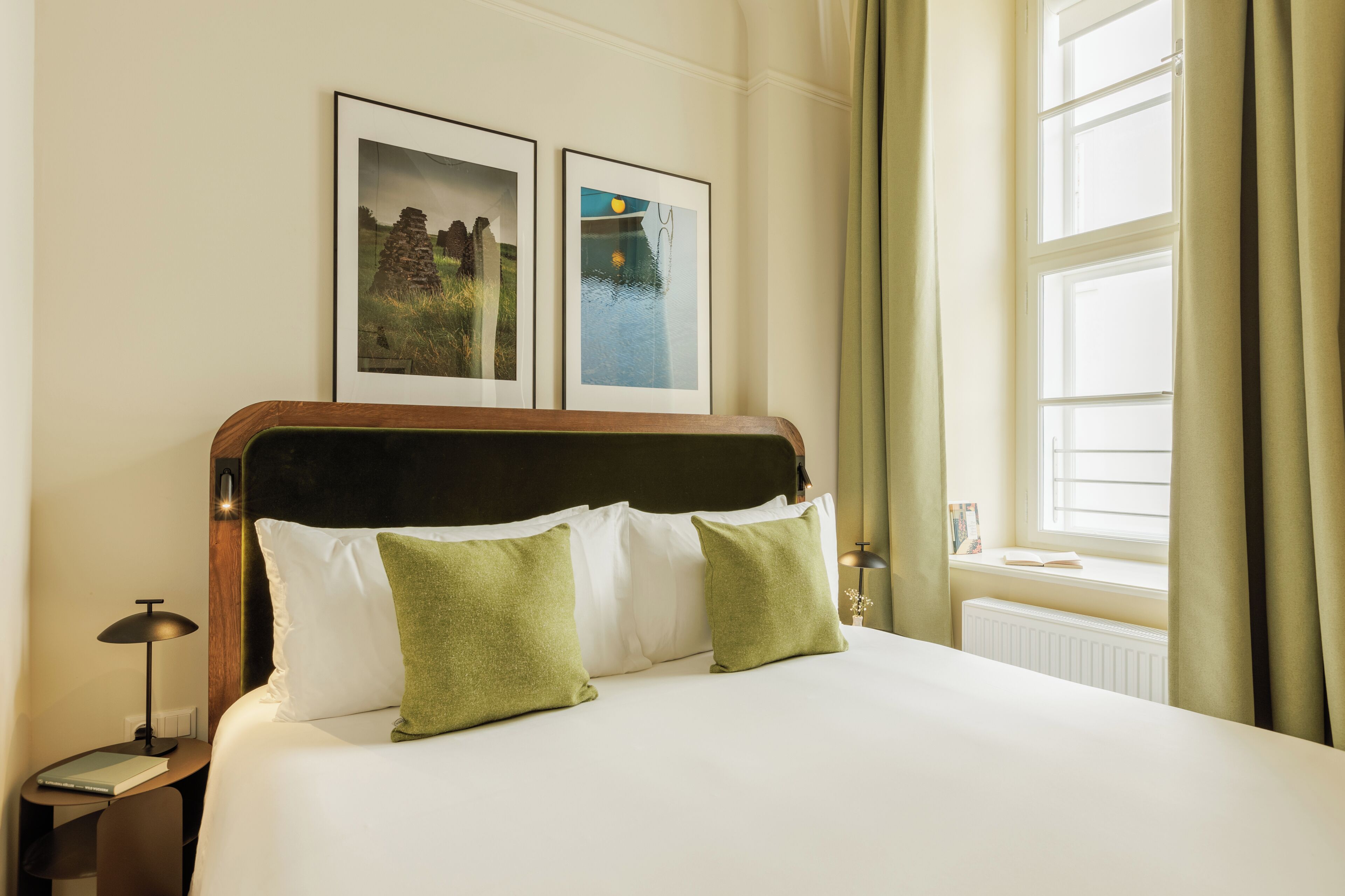 Een hotelkamer met een bed, twee ingelijste foto's aan de muur, een nachtkastje met een lamp en een raam met gordijnen.