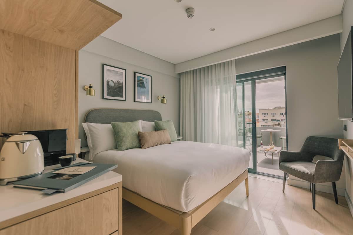 A modern hotel room with a bed, a desk, a chair, and a balcony with a view of buildings.