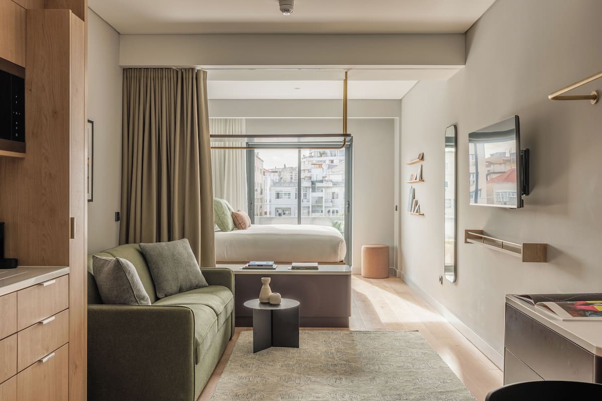 A modern hotel room with a bed, a sofa, a small table, a TV, and a view of the city through large windows.