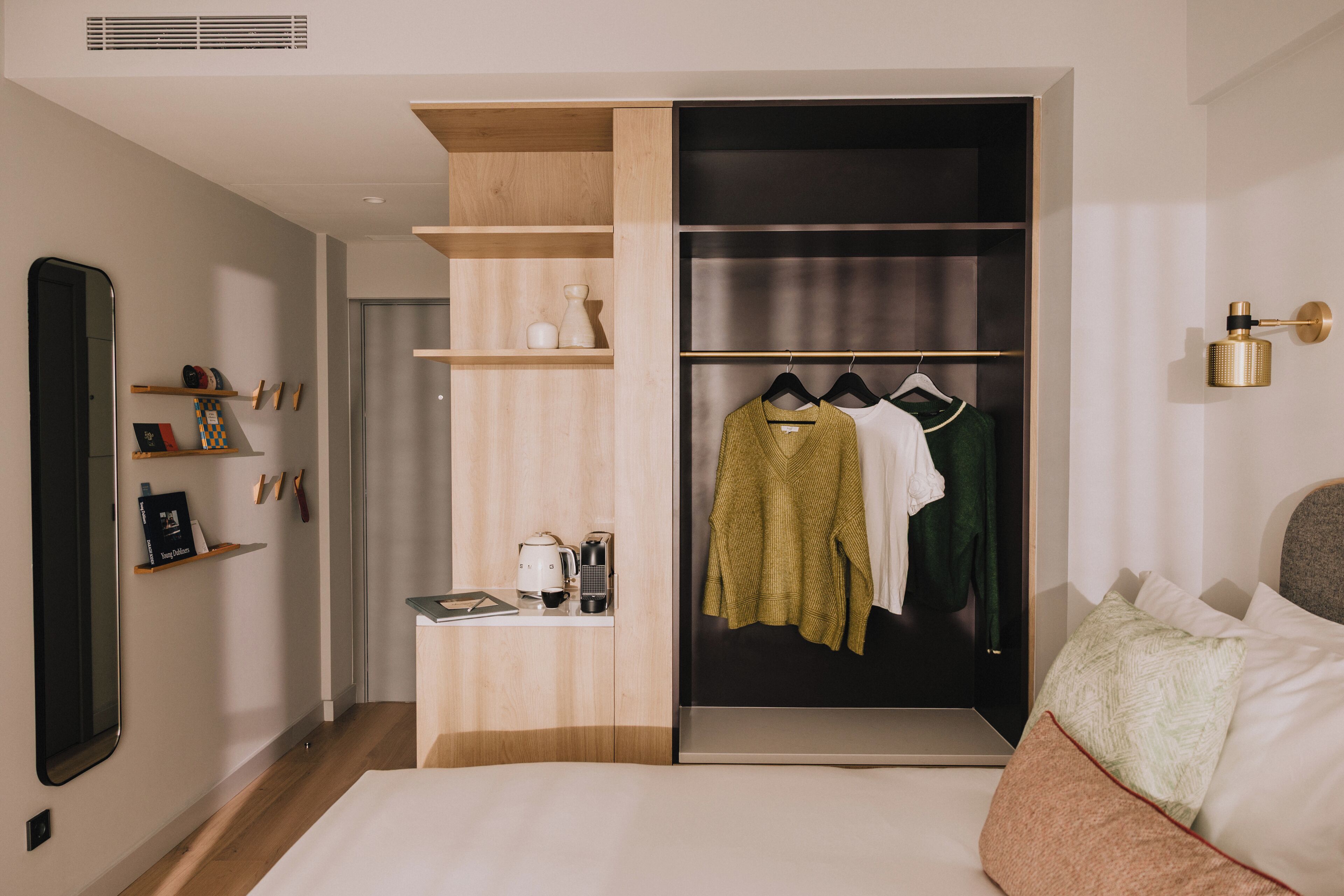 Een moderne slaapkamer met een inbouwkast, een nachtkastje met een boek en een waterkoker, en een bed met kussens.