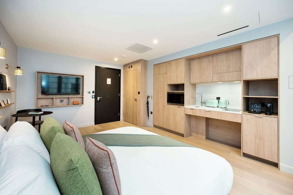 A modern hotel room with a bed, a TV, a kitchenette, and a door leading to the bathroom.