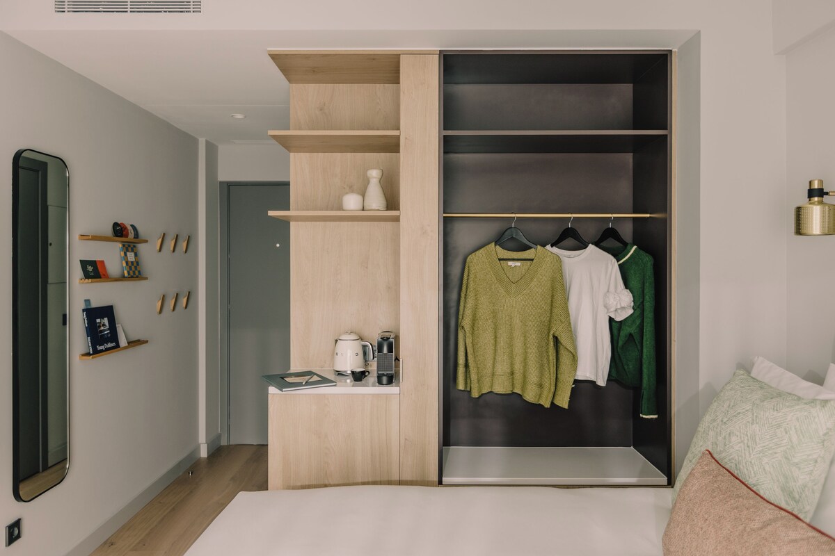 A bedroom with a bed, a wardrobe, a mirror, and a shelf with books and a kettle.