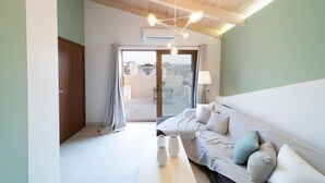 Ein modernes Hotelzimmer mit Holzboden, einem großen Bett mit vielen Kissen, einem weißen Nachttisch mit Vasen und Blick durch eine Glastür nach draußen.
