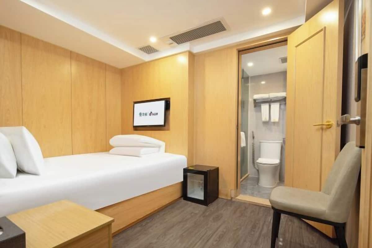 A hotel room with a bed, a small bedside table, a chair, a wooden wardrobe, and a bathroom visible through an open door.