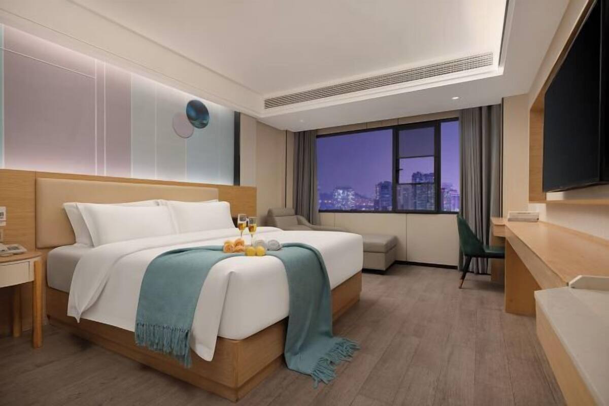 A hotel room with a bed, a desk, a chair, a TV, and a large window with a city view.