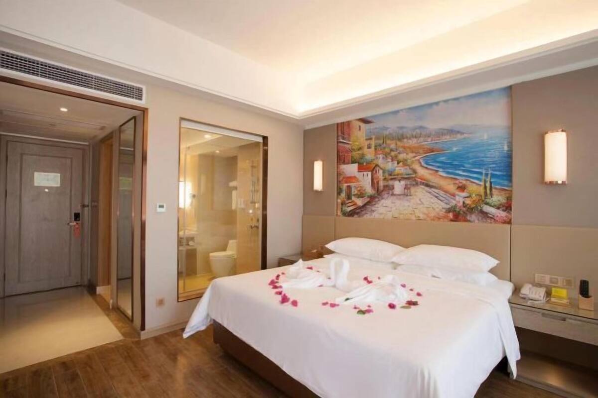 A hotel room with a bed, a painting of a coastal scene, a shower area, and a bedside table with a lamp.