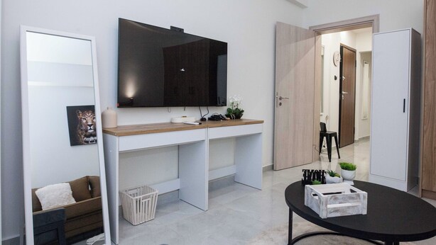 A modern living room with a large flat-screen TV mounted on the wall, a wooden console table, a black coffee table, and a white sofa. - Brand New 1 Bd Apt in Trendy Kipseli (Athens)