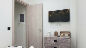 A hotel room with a bed, a wooden wardrobe, a television mounted on the wall, and a door leading to another room. - Brand New 1 Bd Apt in Trendy Kipseli (Athens)
