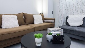 A modern living room with a brown sofa, white fluffy pillows, a black coffee table with plants and decorative items, and a grey sofa with a white throw. - Brand New 1 Bd Apt in Trendy Kipseli (Athens)
